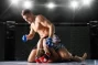 An intense moment in an MMA fight as one fighter performs a ground and pound move on his opponent inside the cage, with bright lights illuminating the arena.